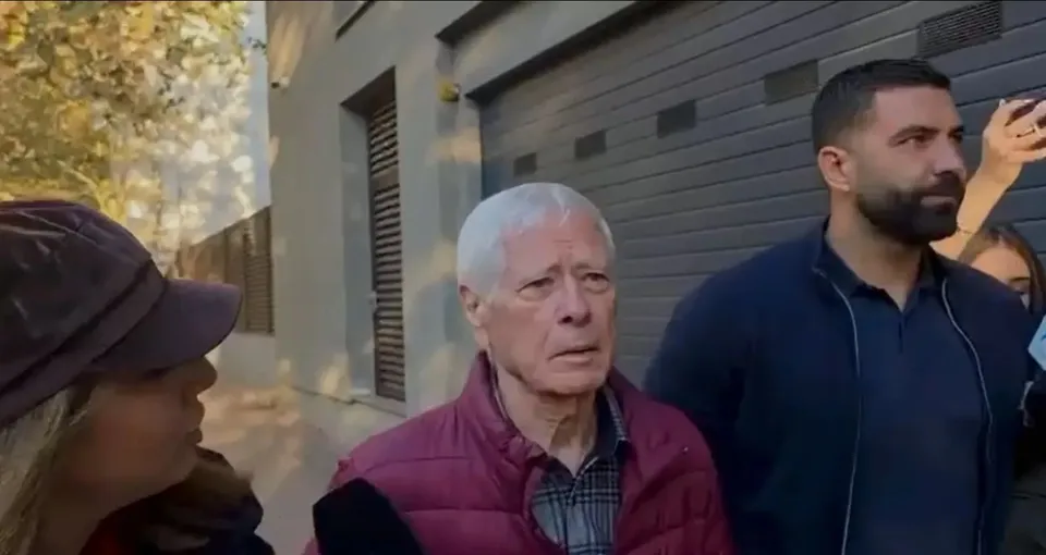 Un bărbat cu părul alb și o jachetă roșie este încadrat între o femeie cu șapcă și un bărbat cu barbă purtând un hanorac albastru.