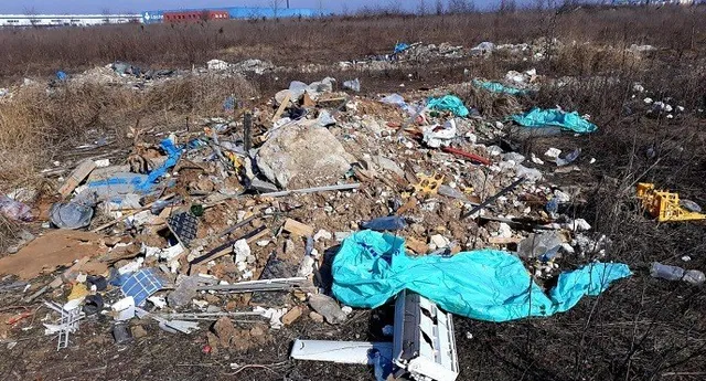Un morman de deșeuri diverse, inclusiv obiecte din plastic și resturi metalice, este împrăștiat pe un teren cu vegetație uscată sub cerul senin.
