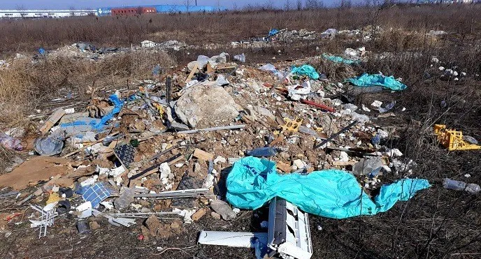 Un morman de deșeuri diverse, inclusiv obiecte din plastic și resturi metalice, este împrăștiat pe un teren cu vegetație uscată sub cerul senin.