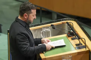 Un bărbat cu barbă și păr scurt, îmbrăcat în sacou negru, stă în fața unui podium de lemn și citește un document alb.