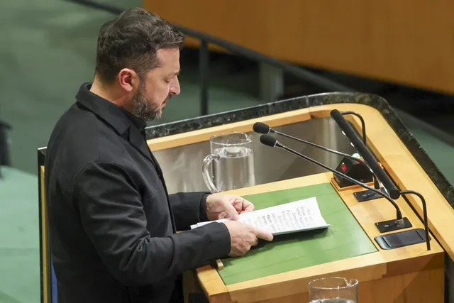 Un bărbat cu barbă și păr scurt, îmbrăcat în sacou negru, stă în fața unui podium de lemn și citește un document alb.