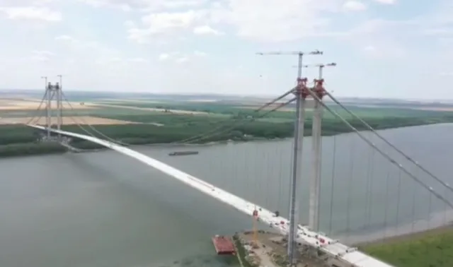 O vedere aeriană a unui pod suspendat lung care traversează un fluviu larg sub un cer parțial înzăpezit.