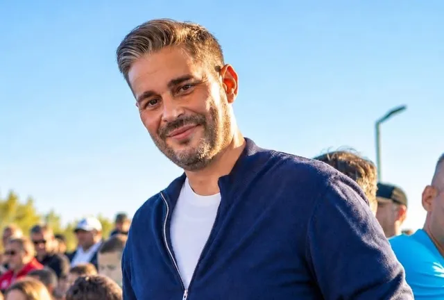 Un bărbat cu părul scurt și gri, purtând un tricou alb și un hanorac albastru, zâmbește în fața unui cer senin și a unor persoane.