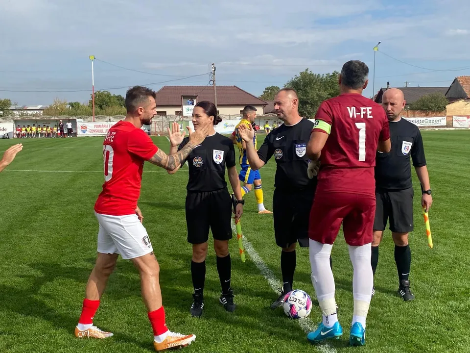Cinci persoane îmbrăcate în echipamente sportive și de arbitraj stau pe un teren de fotbal verde sub un cer senin.