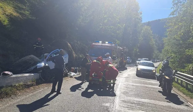 O echipă de salvatori în uniforme colorate lucrează pe un drum asfaltat lângă un autoturism alb și un vehicul de serviciu în mijlocul pădurii.
