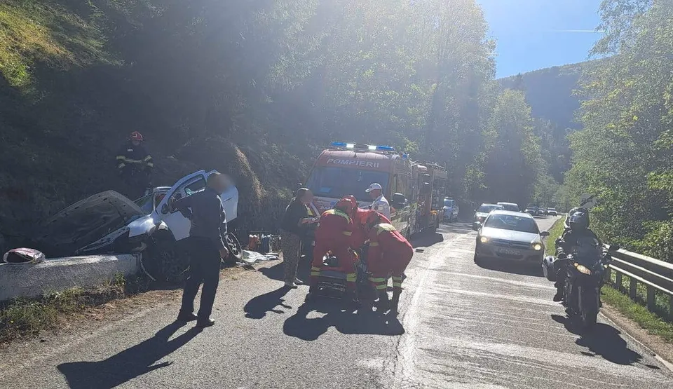 O echipă de salvatori în uniforme colorate lucrează pe un drum asfaltat lângă un autoturism alb și un vehicul de serviciu în mijlocul pădurii.