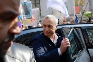 Un bărbat cu părul scurt și alb, îmbrăcat într-o jachetă închisă la culoare, stă lângă o mașină deschisă lângă un grup de oameni.
