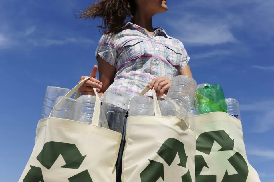 O persoană poartă două sacoșe de pânză cu simboluri de reciclare, pline cu sticle de plastic transparente și una verde, sub un cer albastru.
