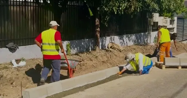 Trei muncitori cu veste reflectorizante lucrează pe un șantier de pavaj lângă un bord curb, având un cărucior roșu și materiale de construcție în apropiere.