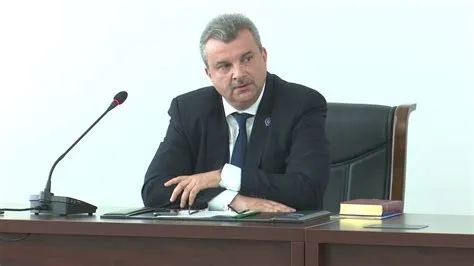 Un bărbat cu mustață, îmbrăcat în costum închis și cravată, stă la o masă de lemn lângă un microfon și un dosar.