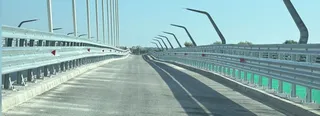 O porțiune de drum asfaltat este delimitată de bariere metalice și stâlpi de iluminat pe un pod sub un cer senin.