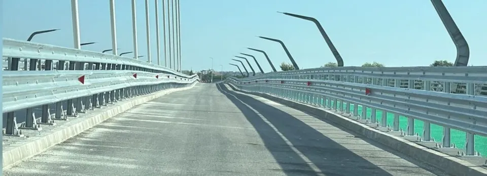 O porțiune de drum asfaltat este delimitată de bariere metalice și stâlpi de iluminat pe un pod sub un cer senin.