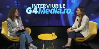 Două femei stau așezate în fotolii galbene, discutând într-un studio cu un fundal albastru pe care scrie un text alb și albastru.