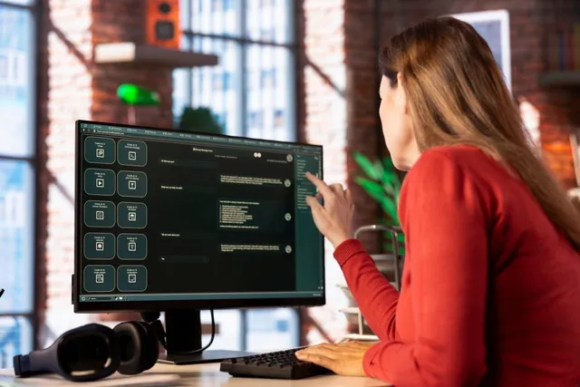 O femeie cu părul lung și bluză roșie stă la un birou și indică un monitor de calculator care afișează diverse pictograme și text.