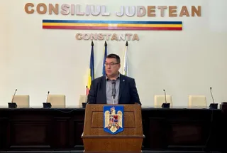 Un bărbat cu ochelari și sacou închis stă în spatele unui podium de lemn în fața unui tabel cu microfoane și un perete alb.