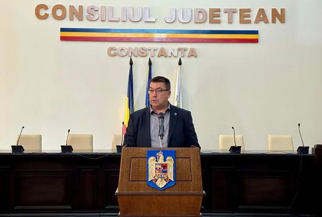 Un bărbat cu ochelari și sacou închis stă în spatele unui podium de lemn în fața unui tabel cu microfoane și un perete alb.