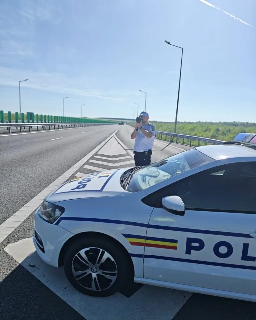 Un agent de poliție stă pe marginea unei autostrăzi lângă o mașină de poliție albă sub un cer senin.