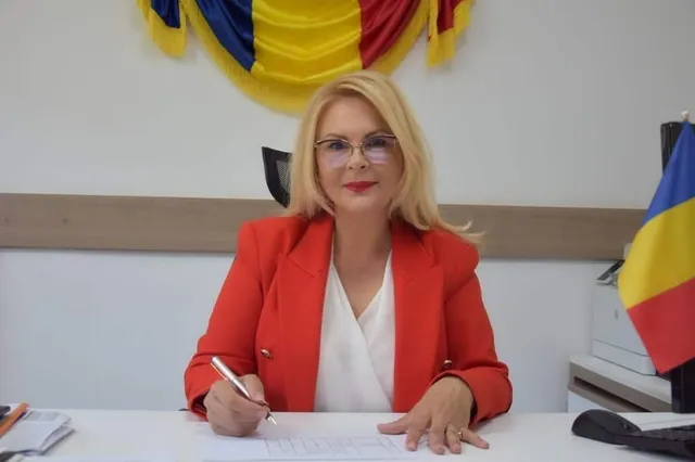 O femeie cu părul blond și ochelari, purtând un sacou roșu peste o bluză albă, stă la o masă cu un pix în mână.