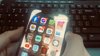 O persoană ține un smartphone în mâini, afișând diverse aplicații de rețele sociale și comunicare pe ecranul activ, în fața unui tastatură de calculator.