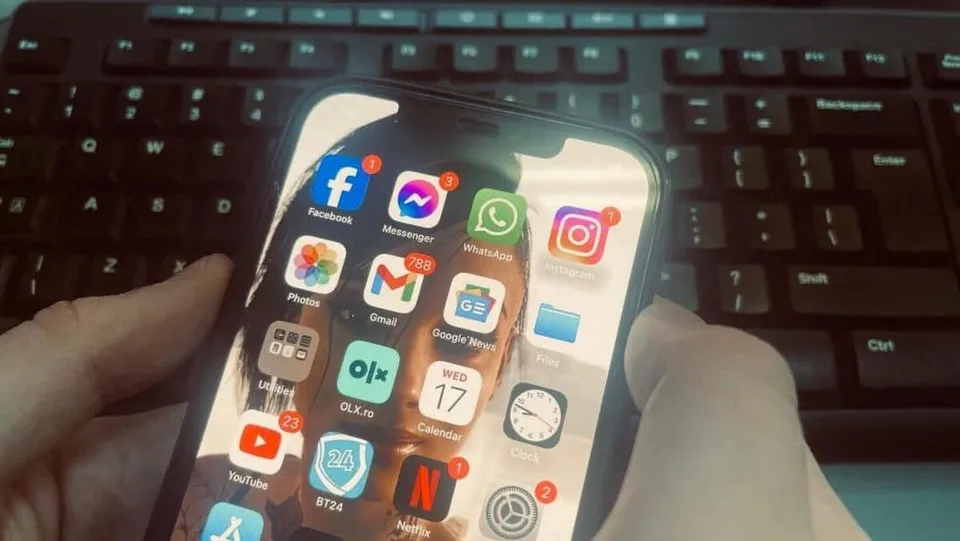 O persoană ține un smartphone în mâini, afișând diverse aplicații de rețele sociale și comunicare pe ecranul activ, în fața unui tastatură de calculator.