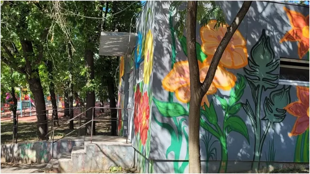 O clădire mică cu picturi murale florale colorate este situată lângă copaci într-o zonă cu vegetație și o rampă de acces metalică.