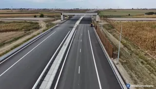 O autostradă cu două caraje separate de o barieră centrală traversează un peisaj agricol sub un cer înnorat.