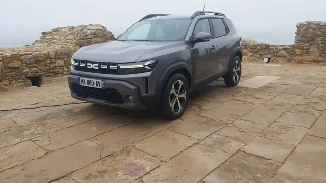 Un autoturism gri este parcat pe o suprafață pavată lângă un zid de piatră sub un cer înnorat.