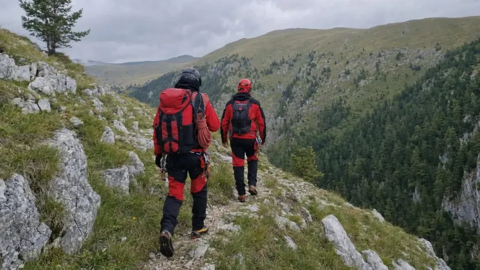 Două persoane purtând rucsacuri și echipament de munte merg pe un potecă îngustă de pe un versant muntos stâncos.