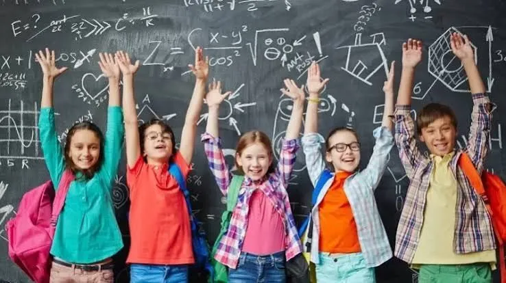 Șase copii cu brațele ridicate stau în fața unei table negre pe care sunt scrise formule matematice și desene cu cretă albă.