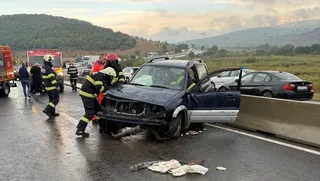 Pompieri în echipament de protecție cu benzi reflectorizante lucrează lângă un autoturism de culoare închisă avariat, situat lângă o barieră metalică de protecție pe un drum umed.