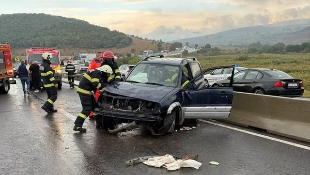 Pompieri în echipament de protecție cu benzi reflectorizante lucrează lângă un autoturism de culoare închisă avariat, situat lângă o barieră metalică de protecție pe un drum umed.
