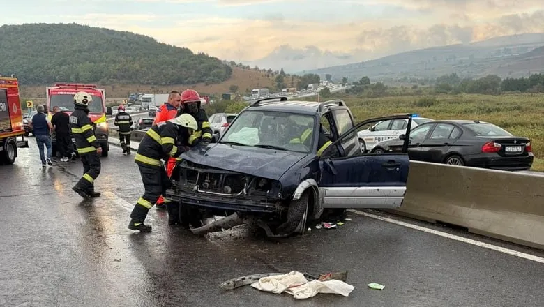 Pompieri în echipament de protecție cu benzi reflectorizante lucrează lângă un autoturism de culoare închisă avariat, situat lângă o barieră metalică de protecție pe un drum umed.