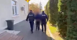 Trei persoane purtând uniformă închisă cu inscripția POLITIA merg pe un trotuar lângă un zid alb și un gard de vegetație înaltă.