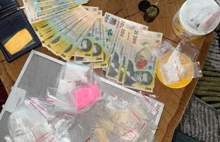 Multe bancnote de lei și diverse pungi mici de plastic conținând substanțe albe sau colorate sunt întinse pe o suprafață de lemn și metal.