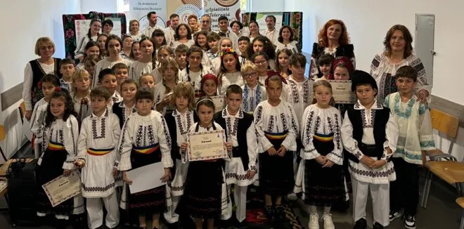 Un grup de copii și adulți poartă cămăși albe cu broderii și stau în grup într-o încăpere cu un perete alb în fundal.