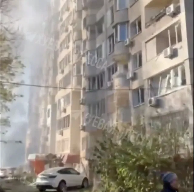 Un bloc de apartamente înalt este situat lângă un drum unde o mașină albă este parcată lângă vegetație.