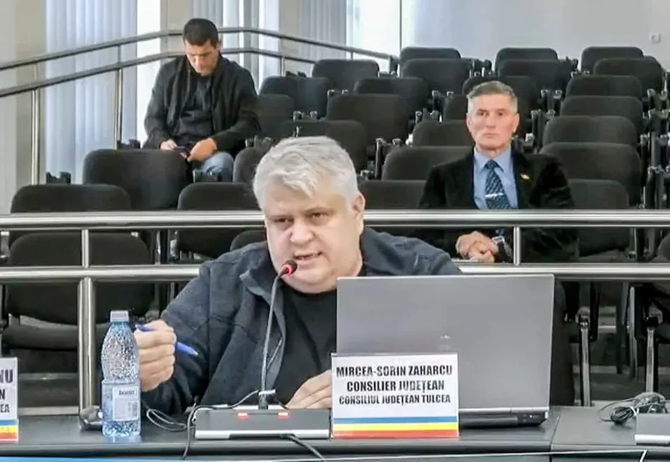 Un bărbat cu părul alb stă la o masă cu un laptop, un microfon și o sticlă de apă în fața sa.