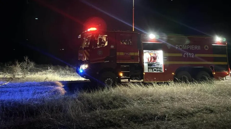 Un autospecial de pompieri roșu cu marcajul POMPIERII este parcat pe un teren cu iarbă uscată sub lumină nocturnă.