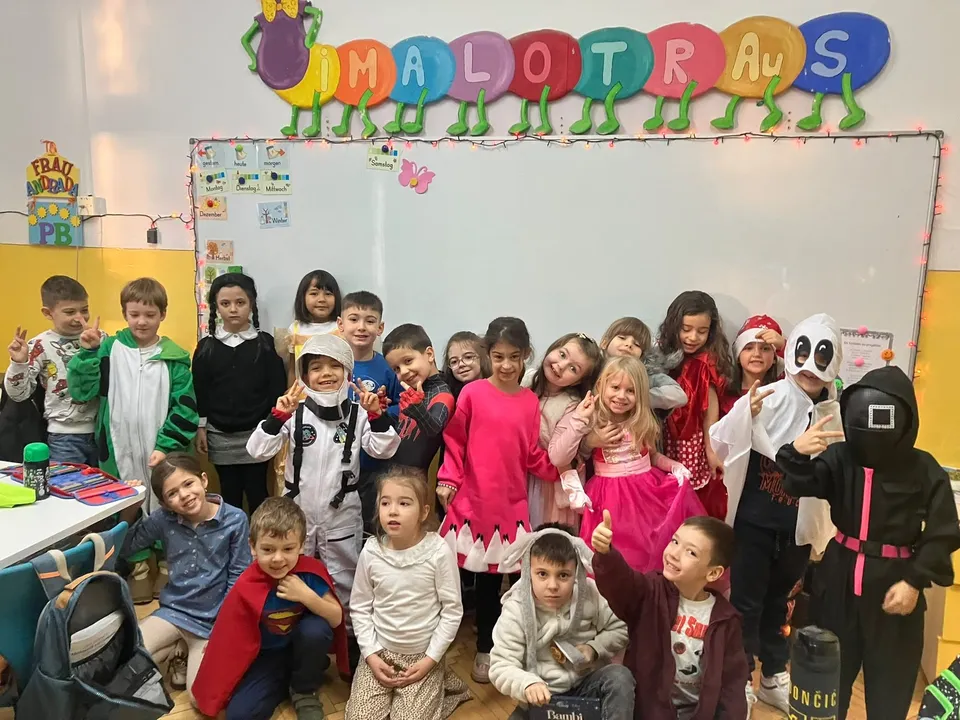 Un grup de copii îmbrăcați în costume de carnaval stau în fața unui panou alb într-o sală de clasă decorată colorat.