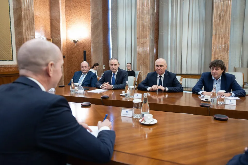 Cinci bărbați în costume purtând cămăși și cravate stau la o masă lungă de lemn într-o încăpere cu pereți din marmură.