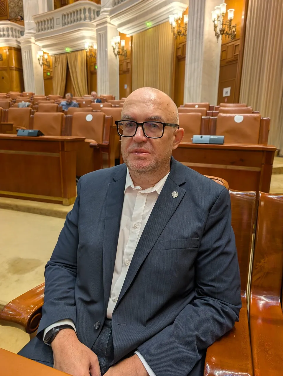 Un bărbat cu ochelari și sacou închis la culoare stă așezat pe un scaun de piele într-o sală cu birouri de lemn și coloane albe.