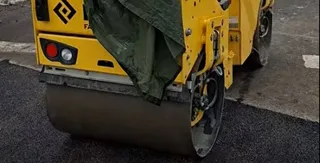 Un aparat de compactat galben cu un cilindru metalic este poziționat pe o suprafață de asfalt proaspăt întinsă.