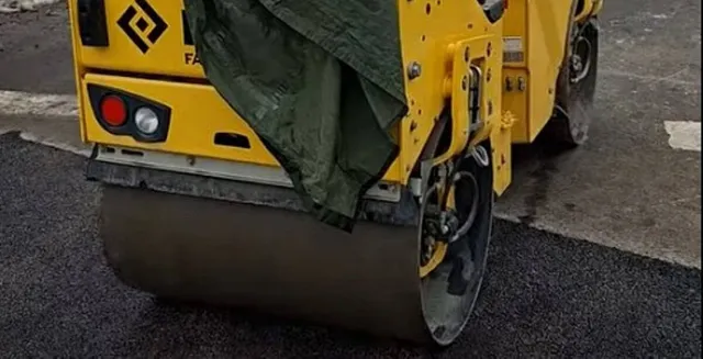 Un aparat de compactat galben cu un cilindru metalic este poziționat pe o suprafață de asfalt proaspăt întinsă.