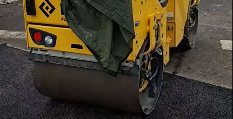 Un aparat de compactat galben cu un cilindru metalic este poziționat pe o suprafață de asfalt proaspăt întinsă.