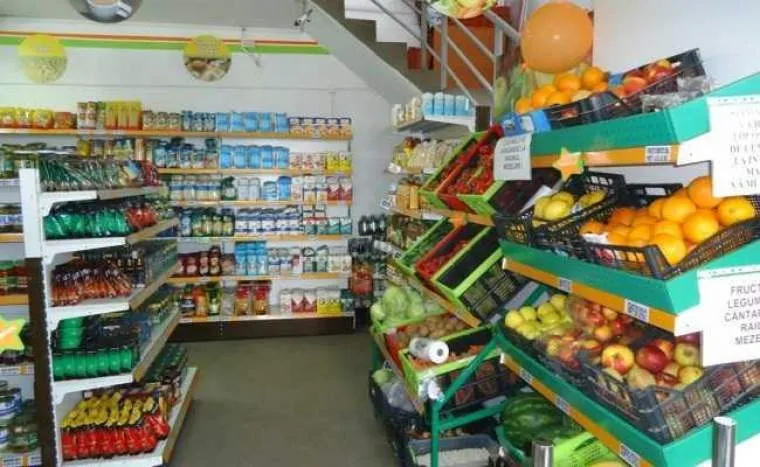 Rafturile unui magazin sunt umplute cu diverse produse alimentare, inclusiv fructe și legume așezate în coșuri pe suporturi de culoare verde.