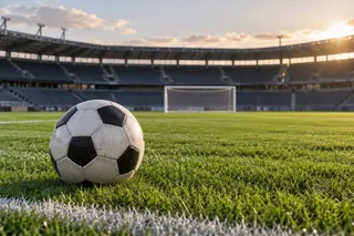 Un fotbal alb și negru este situat pe iarba unui stadion de fotbal, cu tribunele și un por în fundal.