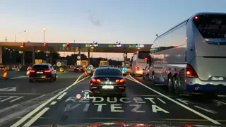 Mașini și autobuze circulă pe o autostradă spre o barieră de taxare sub un cer de seară.