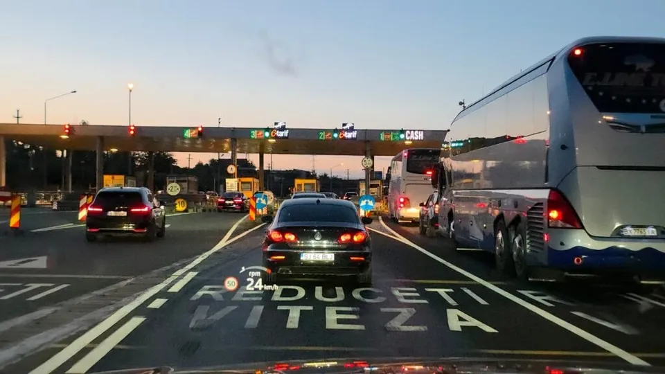 Mașini și autobuze circulă pe o autostradă spre o barieră de taxare sub un cer de seară.