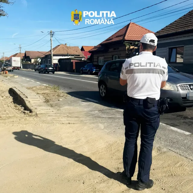 Un polițist cu tricou alb și pantaloni închiși stă pe marginea unui drum lângă o mașină gri sub un cer senin.