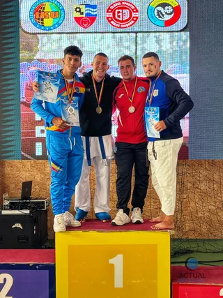Patru bărbați stau pe un podium, purtând costume de karate și medalii, în fața unui ecran mare cu diverse embleme colorate.